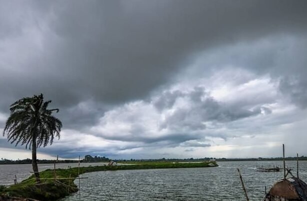 Dramatic monsoon clouds gather over a serene rural landscape featuring a river or lake, a lone palm tree leaning on the edge of an eroded riverbank, and a small thatched fishing hut on stilts in the water. The dark clouds dominate the sky, indicating an impending storm, while the water appears choppy and the greenery in the foreground adds contrast to the moody atmosphere.